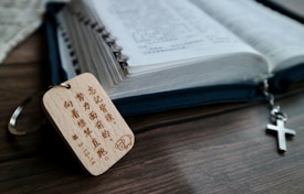 An open book with text in an Asian script lies on a wooden table. Next to it is a keychain with wooden tag engraved with text, and a metal cross hanging as part of the zipper pull.