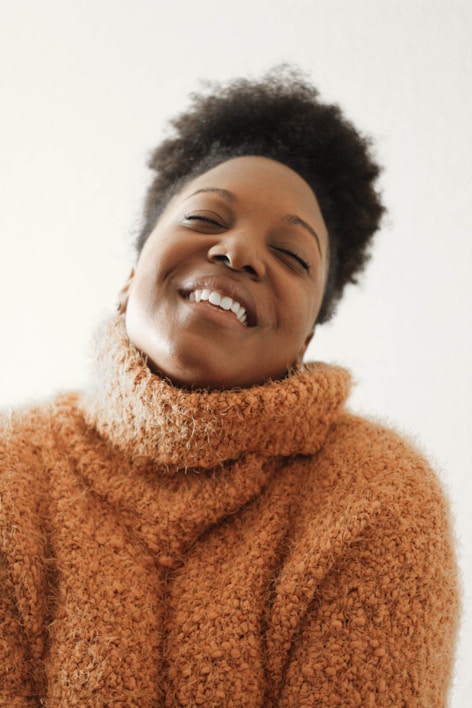 A person with a joyful expression is wearing a chunky, textured sweater. The sweater is a warm, cozy knit, and the person's eyes are closed with a content smile showing teeth. The background is a plain, light color, enhancing the warmth and comfort conveyed by the scene.