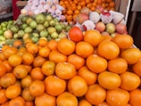 A vibrant display of various fruits including a large pile of oranges in the foreground, surrounded by apples wrapped in protective netting, ripe guavas with some leaves attached, and several other fruits including green limes. The arrangement is colorful and neatly organized, likely from a market or fruit stall.