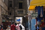 A narrow alleyway lined with worn, rustic buildings. On the left, two people walk together, carrying backpacks. To the right, a small shop displays colorful hanging bags under a yellow awning. A few men are seated or standing nearby, adding to the bustling atmosphere. A weathered sign directs to a restaurant and tea shop.