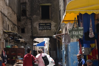 A narrow alleyway lined with worn, rustic buildings. On the left, two people walk together, carrying backpacks. To the right, a small shop displays colorful hanging bags under a yellow awning. A few men are seated or standing nearby, adding to the bustling atmosphere. A weathered sign directs to a restaurant and tea shop.