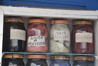 Four glass jars are displayed on shelves, each filled with different colored powders. Labels on the jars indicate 'OCRE', 'ROUGE de Marrakech', 'Vert Noyer', and 'Coquelicot'. The jars have brown lids and appear to be part of a traditional market or shop setting.