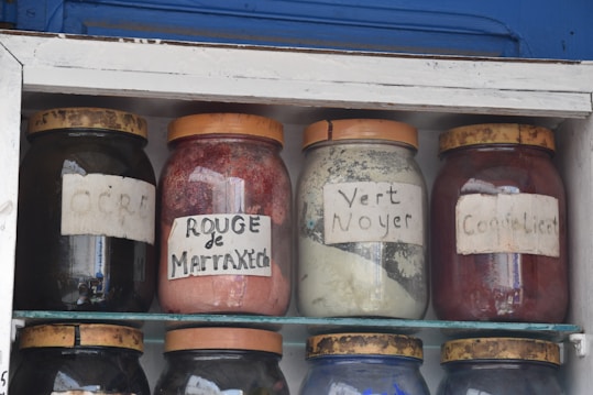 Four glass jars are displayed on shelves, each filled with different colored powders. Labels on the jars indicate 'OCRE', 'ROUGE de Marrakech', 'Vert Noyer', and 'Coquelicot'. The jars have brown lids and appear to be part of a traditional market or shop setting.