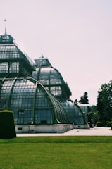 An intricate glass and metal greenhouse structure stands prominently, reflecting light and casting shadows. The surrounding area includes neatly trimmed grass and some trees, suggesting a garden or park setting. The architectural detail of the greenhouse includes curves and a combination of opaque and transparent panes.