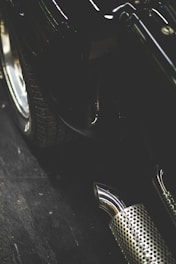 A close-up view of an automobile's tire and exhaust pipe. The image includes a focus on the metallic, perforated casing of the exhaust, as well as the tread of the tire and parts of the vehicle's bodywork.