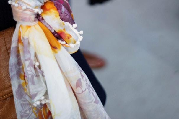 A close-up view of a colorful scarf with patterns in orange, purple, and yellow hues, featuring small white pom-poms along the edge. The scarf is draped over a textured brown leather surface with part of a darker fabric visible in the background.
