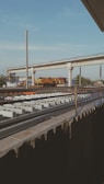 A railway track under construction is visible in the foreground, with rows of concrete sleepers laid out in preparation for the tracks. Above this, an elevated railway or bridge is partially developed, supported by concrete columns. A yellow and black train or maintenance vehicle is parked on the bridge, surrounded by construction equipment and materials. The sky is mostly clear with a few clouds, and there are trees and buildings in the background.
