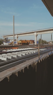 A railway track under construction is visible in the foreground, with rows of concrete sleepers laid out in preparation for the tracks. Above this, an elevated railway or bridge is partially developed, supported by concrete columns. A yellow and black train or maintenance vehicle is parked on the bridge, surrounded by construction equipment and materials. The sky is mostly clear with a few clouds, and there are trees and buildings in the background.