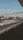 A railway track under construction is visible in the foreground, with rows of concrete sleepers laid out in preparation for the tracks. Above this, an elevated railway or bridge is partially developed, supported by concrete columns. A yellow and black train or maintenance vehicle is parked on the bridge, surrounded by construction equipment and materials. The sky is mostly clear with a few clouds, and there are trees and buildings in the background.