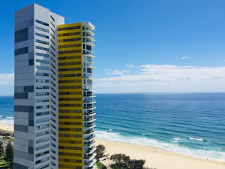 A modern high-rise building with a contrasting color scheme of gray and yellow stands adjacent to a calm, expansive beach. The ocean stretches out under a clear blue sky with a few scattered clouds. Sparse greenery and trees are visible near the sandy shoreline.