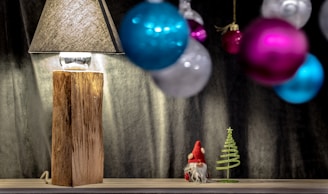 A rustic wooden lamp with a textured shade sits on a wooden surface. To the side, there's a small figurine resembling a gnome next to a decorative metal Christmas tree. In the foreground, blurred colorful baubles in shades of pink, blue, and silver hang, adding to the festive atmosphere.