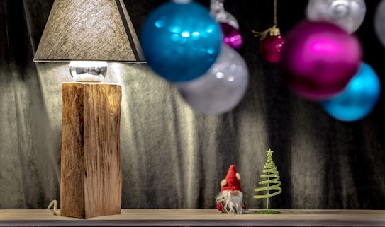A rustic wooden lamp with a textured shade sits on a wooden surface. To the side, there's a small figurine resembling a gnome next to a decorative metal Christmas tree. In the foreground, blurred colorful baubles in shades of pink, blue, and silver hang, adding to the festive atmosphere.