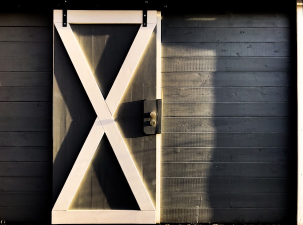 A wooden door with a prominent white X-shaped design stands out against a wall of horizontal dark wooden planks. The texture of the wood is visible, and soft light casts shadows on the surface, adding depth and contrast.
