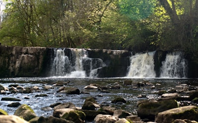 A serene natural landscape featuring a waterfall cascading over rocky ledges, surrounded by lush green trees and vegetation. The water flows into a calm river with stones scattered across the surface, creating a peaceful and tranquil atmosphere.