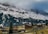 A mountainous landscape features snow-covered peaks partly obscured by clouds. Below the mountains, a small village with traditional wooden houses and scattered trees sits on a gently sloped hillside. The scene is serene with muted colors and hints of greenery.