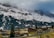 A mountainous landscape features snow-covered peaks partly obscured by clouds. Below the mountains, a small village with traditional wooden houses and scattered trees sits on a gently sloped hillside. The scene is serene with muted colors and hints of greenery.