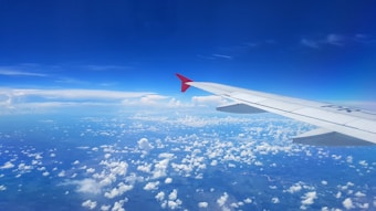 The view from an airplane window shows a clear blue sky and scattered white clouds below. The aircraft's wing, with a red tip, extends diagonally across the image, suggesting a sense of travel and flight.