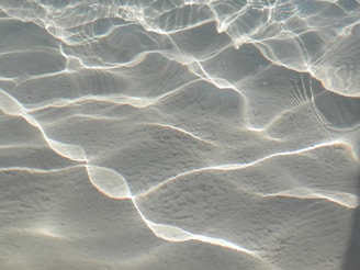 Waves of light are refracted and reflected on a sandy underwater surface, creating intricate patterns of light and shadow. The gentle play of light gives an ethereal and calm appearance.
