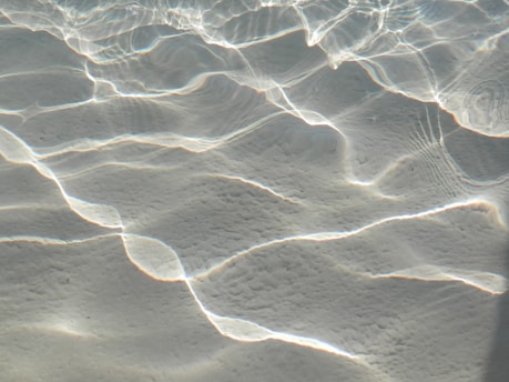 Waves of light are refracted and reflected on a sandy underwater surface, creating intricate patterns of light and shadow. The gentle play of light gives an ethereal and calm appearance.