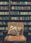 A cozy library setting featuring a cushioned armchair with a decorative pillow placed in front of bookshelves filled with various books. The shelves are packed with colorful book spines, creating a warm and inviting atmosphere.
