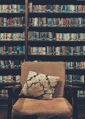 A cozy library setting featuring a cushioned armchair with a decorative pillow placed in front of bookshelves filled with various books. The shelves are packed with colorful book spines, creating a warm and inviting atmosphere.
