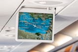 An in-flight entertainment screen displays a digital map showing the current location of a plane flying over the Mediterranean region. The map includes cities such as Athens, Beirut, Tel Aviv, and Cairo. The airplane icon indicates the flight path, with blue representing the sea and green for the land. The screen is mounted in an overhead compartment of the airplane, with visible cabin lights in the background.