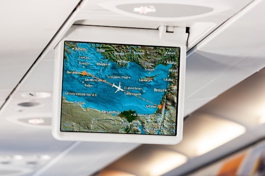 An in-flight entertainment screen displays a digital map showing the current location of a plane flying over the Mediterranean region. The map includes cities such as Athens, Beirut, Tel Aviv, and Cairo. The airplane icon indicates the flight path, with blue representing the sea and green for the land. The screen is mounted in an overhead compartment of the airplane, with visible cabin lights in the background.
