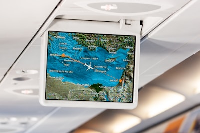 An in-flight entertainment screen displays a digital map showing the current location of a plane flying over the Mediterranean region. The map includes cities such as Athens, Beirut, Tel Aviv, and Cairo. The airplane icon indicates the flight path, with blue representing the sea and green for the land. The screen is mounted in an overhead compartment of the airplane, with visible cabin lights in the background.