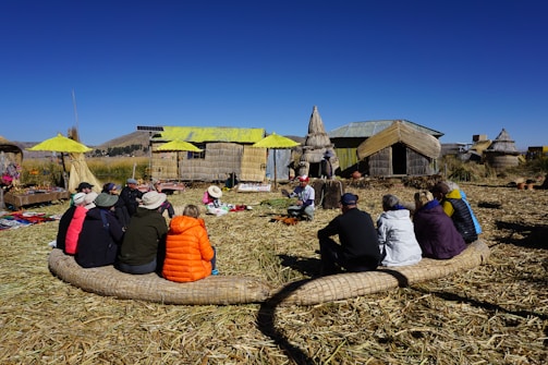 A group of people seated in a circle on a grassy area, attentively listening to a person in traditional attire. They are surrounded by rustic huts made of reeds with a bright blue sky in the background. The setting appears to be a village or cultural site, with handmade crafts displayed nearby.
