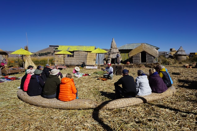 A group of people seated in a circle on a grassy area, attentively listening to a person in traditional attire. They are surrounded by rustic huts made of reeds with a bright blue sky in the background. The setting appears to be a village or cultural site, with handmade crafts displayed nearby.