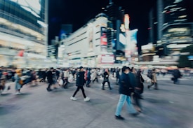A bustling city scene at night with many people crossing a street surrounded by tall buildings adorned with bright lights and digital billboards. The image captures a sense of motion and energy with a slight blur effect, indicating movement.