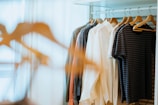 A closet filled with various clothing items hanging on wooden hangers. The clothing includes shirts, jackets, and tops in a range of colors such as white, grey, and black. The lighting is soft, with sunlight filtering through a sheer curtain in the background, creating a calm and organized atmosphere.