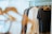 A closet filled with various clothing items hanging on wooden hangers. The clothing includes shirts, jackets, and tops in a range of colors such as white, grey, and black. The lighting is soft, with sunlight filtering through a sheer curtain in the background, creating a calm and organized atmosphere.