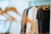 A closet filled with various clothing items hanging on wooden hangers. The clothing includes shirts, jackets, and tops in a range of colors such as white, grey, and black. The lighting is soft, with sunlight filtering through a sheer curtain in the background, creating a calm and organized atmosphere.