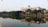 A large, rectangular building with a clock tower on top is situated by a serene lake. The building is adorned with the signage 'VNPT Ha Noi'. Lush green trees line the lakeside, reflecting on the calm water. The sky is clear and light blue, suggesting a tranquil atmosphere.