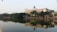 A large, rectangular building with a clock tower on top is situated by a serene lake. The building is adorned with the signage 'VNPT Ha Noi'. Lush green trees line the lakeside, reflecting on the calm water. The sky is clear and light blue, suggesting a tranquil atmosphere.