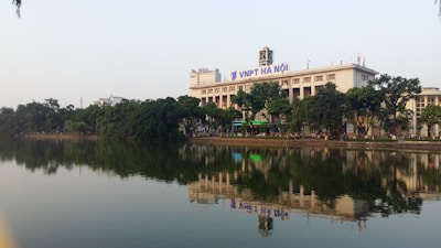 A large, rectangular building with a clock tower on top is situated by a serene lake. The building is adorned with the signage 'VNPT Ha Noi'. Lush green trees line the lakeside, reflecting on the calm water. The sky is clear and light blue, suggesting a tranquil atmosphere.