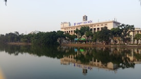A large, rectangular building with a clock tower on top is situated by a serene lake. The building is adorned with the signage 'VNPT Ha Noi'. Lush green trees line the lakeside, reflecting on the calm water. The sky is clear and light blue, suggesting a tranquil atmosphere.