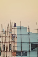 Two people are working on the exterior of a building, seemingly painting or repairing it. The building is surrounded by a framework of bamboo scaffolding. The sky is a soft gradient of pale yellow to orange, suggesting either early morning or late evening. The building itself is painted in light blue and has some decorative elements in darker shades.