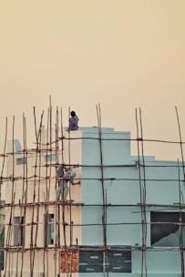 Two people are working on the exterior of a building, seemingly painting or repairing it. The building is surrounded by a framework of bamboo scaffolding. The sky is a soft gradient of pale yellow to orange, suggesting either early morning or late evening. The building itself is painted in light blue and has some decorative elements in darker shades.