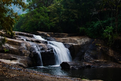 A serene waterfall cascades over smooth, dark rocks surrounded by lush, green forest. The water flows gently into a calm pool below, creating a peaceful natural landscape.
