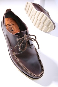 A dark brown leather shoe with detailed stitching and a thick, textured sole is prominently displayed. The shoe features a lace-up design and visible branding on the inner sole. The sole's pattern is visible, showcasing its rugged, slip-resistant design.