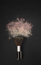 A makeup brush is lying on a dark surface, with a cloud of powder erupting from its bristles. The powder appears fine and has a slightly pinkish hue, spreading out in an artistic pattern.