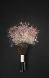 A makeup brush is lying on a dark surface, with a cloud of powder erupting from its bristles. The powder appears fine and has a slightly pinkish hue, spreading out in an artistic pattern.