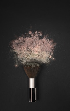 A makeup brush is lying on a dark surface, with a cloud of powder erupting from its bristles. The powder appears fine and has a slightly pinkish hue, spreading out in an artistic pattern.