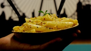 A hand holds a bowl of creamy pasta, garnished with herbs and red chili flakes. Two dark utensils are sticking out of the pasta, and blurred background shows an abstract silhouette of what appears to be a Ferris wheel.
