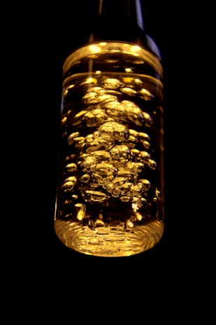 A transparent bottle filled with a golden liquid and illuminated by bright light, showcasing numerous bubbles rising inside. The background is dark, providing a stark contrast that emphasizes the golden hues and clarity of the liquid.
