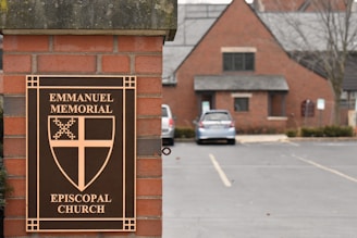 A brick pillar displays a sign for Emmanuel Memorial Episcopal Church. The sign features a decorative cross and shield emblem. In the background, a brick building with a gray roof is visible alongside a parking lot with a few cars.