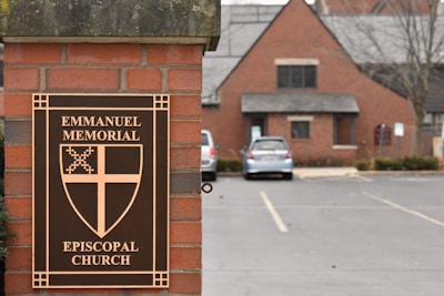 A brick pillar displays a sign for Emmanuel Memorial Episcopal Church. The sign features a decorative cross and shield emblem. In the background, a brick building with a gray roof is visible alongside a parking lot with a few cars.