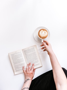A person with a tattoo on the wrist is sitting and holding an open book. The other hand is reaching for a cup of coffee with a heart-shaped latte art on the surface. The background is bright and minimalistic.